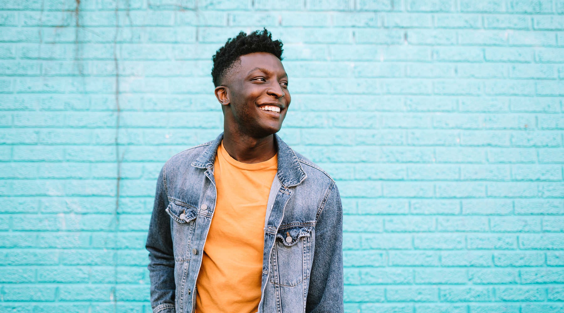 Feel at Home young man in a jeans jacket stands in front of a light blue, brick wall