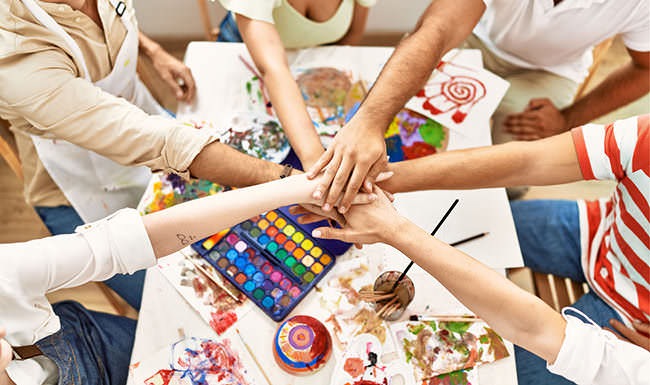 friends place hands over hands in the center over a table strewn with painting supplies